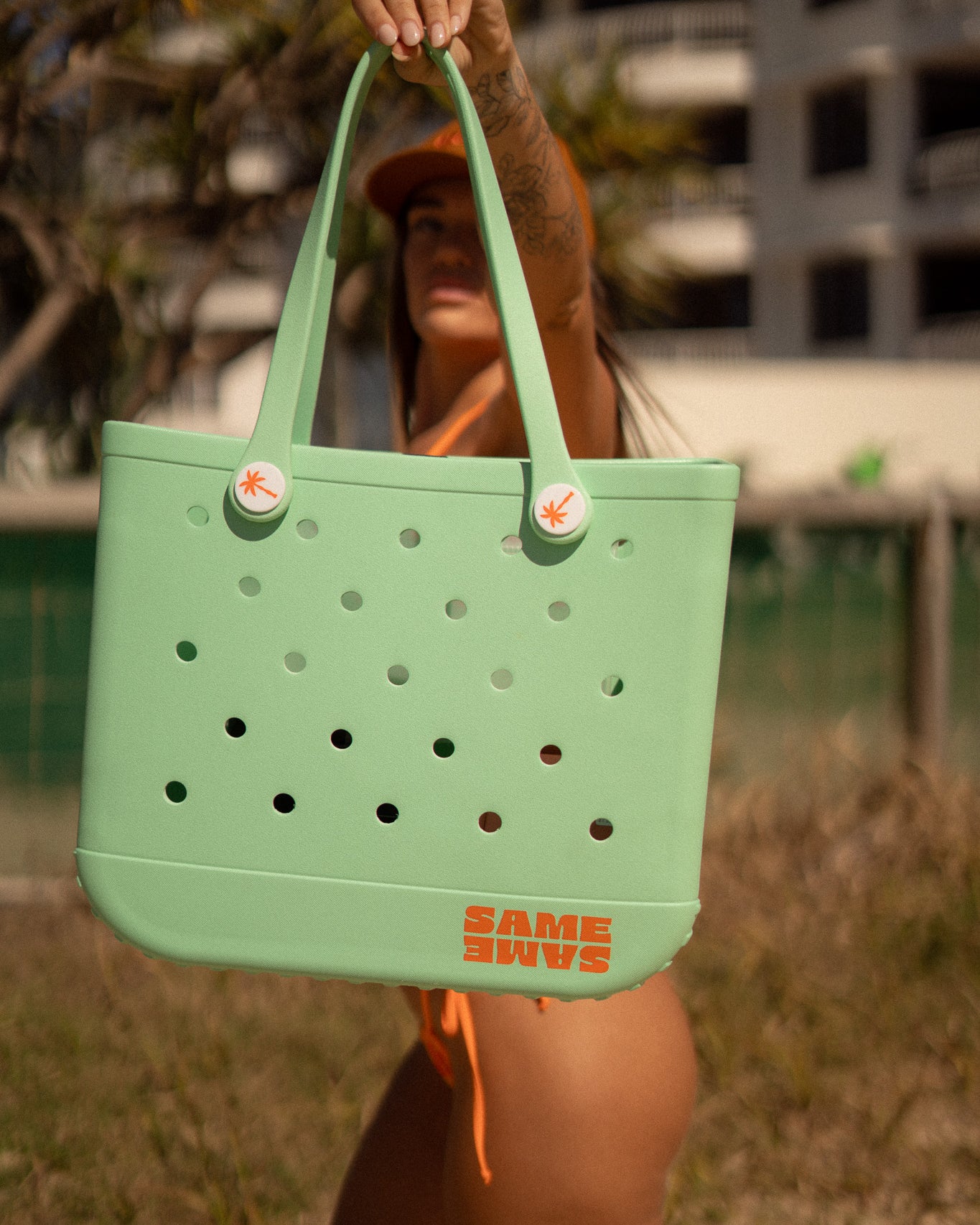 Person holding a green tote bag with 'Same Shins' branding on a beach.