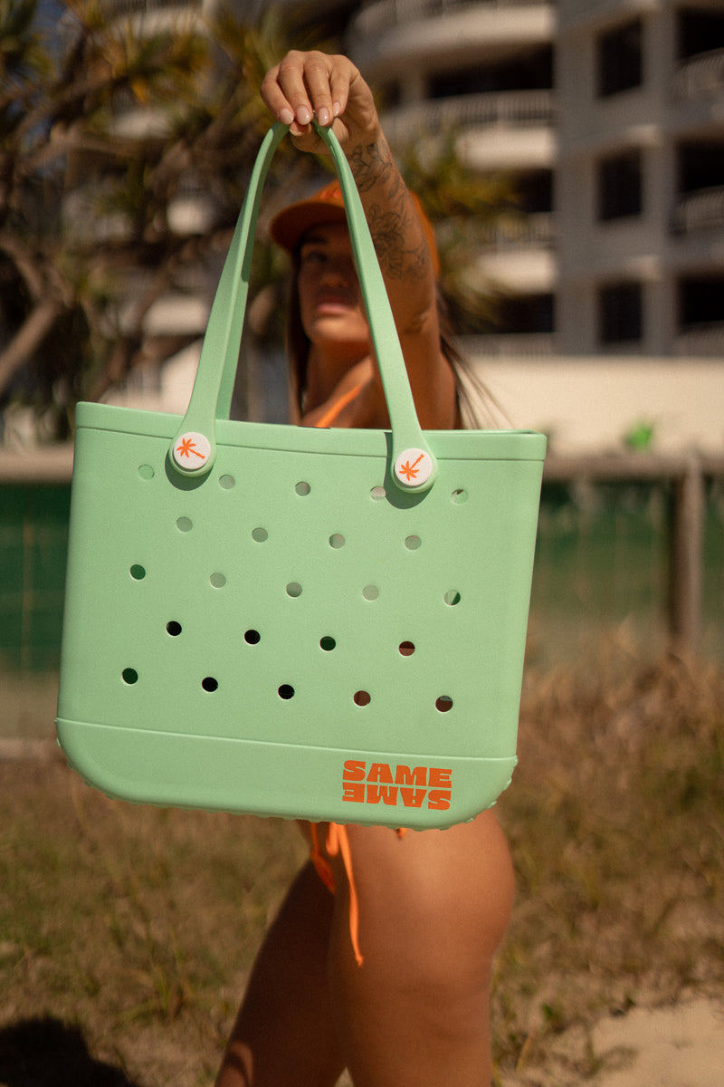 Person holding a green tote bag with 'Same Shins' branding on a beach.